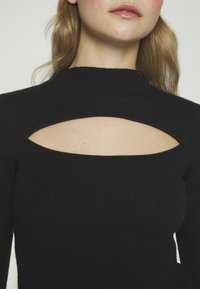 Svart ribbad stickad topp med hög halsringning och utskärning nedanför halsen. Tyget har en mjuk textur och en figurnära design.