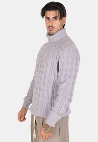 Maglione a punto cable di un delicato colore viola chiaro con collo alto. Presenta bordo e polsini a costine. Morbida trama e vestibilità comoda, indossato da un modello.