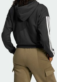 Svart huvtröja sedd bakifrån, med huva och vita ränder längs ärmarna. Sammanförd med olivgröna cargobyxor.