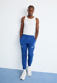 Mand med fletninger iført hvid tanktop, blå joggers med hvide accenter, hvide sokker og klumpede hvide sneakers, der står mod en hvid væg.