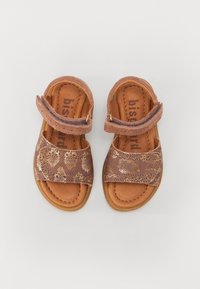 Sandales en cuir marron avec un motif métallique doré, bride à velcro et doublure intérieure beige. Semelle plate et design à bout ouvert.