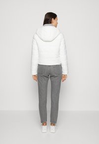 Hvid pufferjakke med hætte, quiltet design og tekstureret overflade. Båret over grå jeans og hvide sneakers, set fra ryggen.