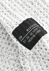 Valge tekstuuriga kangas mustade täpikestega, millel on must silt tekstiga: "LDN MCMLXXXI 51°28'44.8824'' PREMIUM GOODS."
