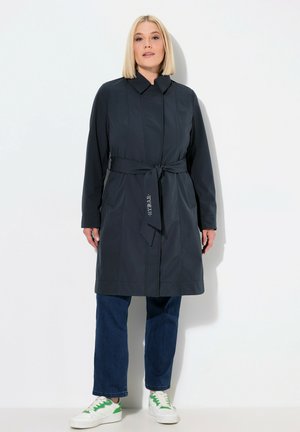 Gabardina azul marino oscura con cinturón, que presenta un cuello, mangas largas y una textura suave. Combinada con vaqueros azules y zapatillas blancas.