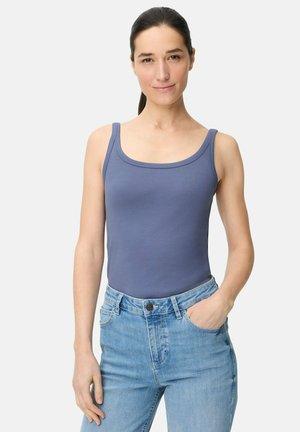 Kobieta o ciemnych włosach związanych z tyłu, ubrana w niebieski top na ramiączkach i jasnoniebieskie dżinsy, stojąca z jedną ręką w kieszeni na jednolitym tle.