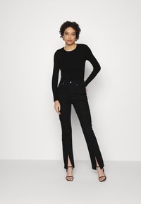 Top preto de mangas longas combinado com jeans pretos de cintura alta com fendas frontais. Modelo usa saltos pretos com tiras. Design simples e ajustado.