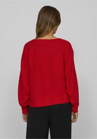 Czerwony sweter z prążkowanego materiału o szerokim dekolcie i bufiastych rękawach, w zestawieniu z czarnymi spodniami. Widok z tyłu ukazuje luźny krój i strukturę.