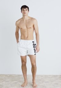 Shorts de bain blancs avec des bandes latérales noires, fabriqués en matériau léger, dotés d'une taille élastique avec un cordon de serrage pour l'ajustement.