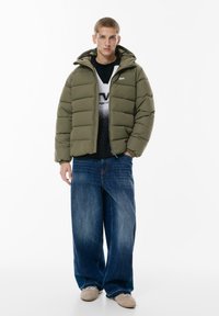 Giacca imbottita verde con cappuccio, chiusura con zip, polsini elasticizzati, indossata sopra una maglietta nera con grafica. Abbinata a jeans a gamba larga blu e scarpe beige.