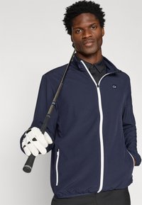 Tengerészkék szélálló dzseki fehér cipzárral, két oldalsó zsebbel és rugalmas mandzsettával. Fekete galléros inggel viselve, és egy golfütőt tartva.