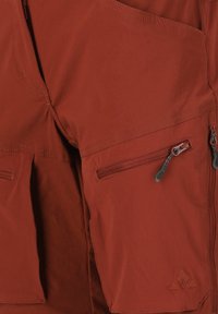 Housut ruosteenvärisessä sävyssä, joissa on useita vetoketjullisia taskuja, teksturoitu kangas ja ommellut yksityiskohdat, sopivat ulkoilma-aktiviteetteihin.