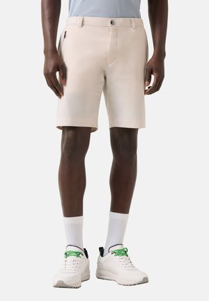 Mann trägt beige Shorts, weiße Socken und weiße Turnschuhe mit grünen Schnürsenkeln, steht vor einem schlichten hellen Hintergrund.