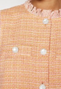 Tweedový kabát v broskvových a růžových tónech s texturovaným povrchem, třepenými okraji a kulatými bílými knoflíky ozdobenými krystaly.