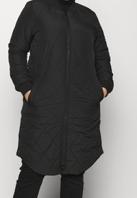 Person som bär en vadderad svart långjacka med dragkedja fram och händerna i sidofickorna mot en enkel bakgrund.