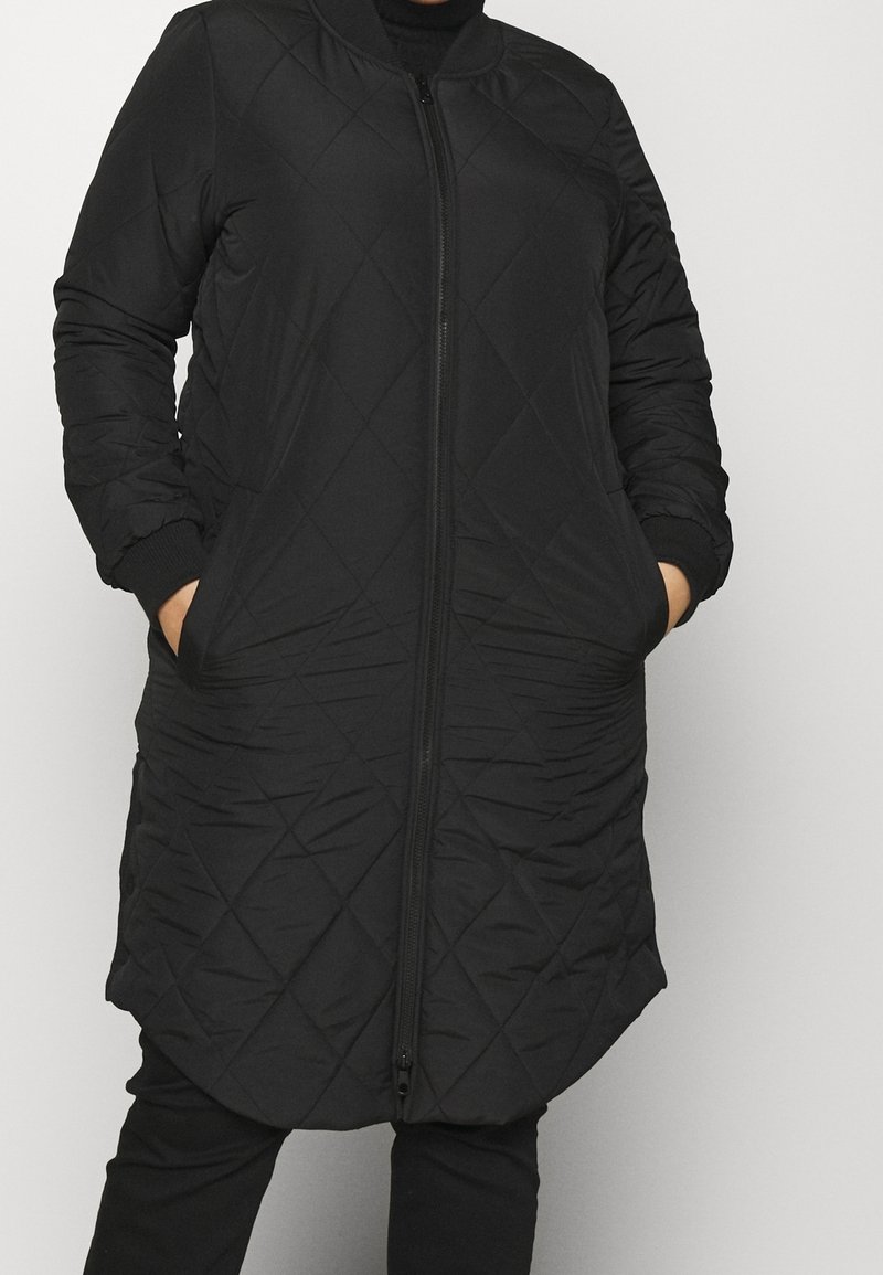 Person som bär en vadderad svart långjacka med dragkedja fram och händerna i sidofickorna mot en enkel bakgrund.