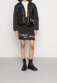 Svart kort puffjacka med glänsande paneler, i kombination med en figursydd svart kjol med vit "Calvin Klein Jeans"-logga. Svarta ankelstövlar.