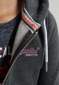 Veste à zip grise avec doublure en polaire, dotée d'un cordon blanc, d'un logo rose brodé et de détails contrastants en gris au niveau du col et de la fermeture éclair.