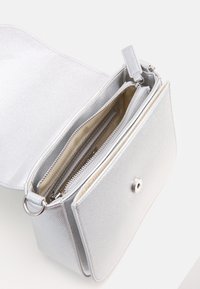Sidabrinė metalinė rankinė su tekstūruota medžiaga, turinti pagrindinį skyrių ir užtrauktuko kišenę, uždarytą užsukimu. Reguliuojama dirželis.