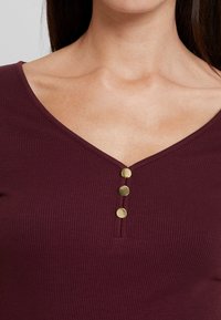 Detalj av en person i en ribbad, mörklila V-ringad tröja med tre runda guldknappar framtill.