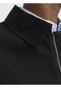 Veste noire avec un col replié, une fermeture éclair élégante et un tissu texturé. Une chemise rayée bleue est visible sous le col.
