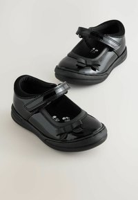 Chaussures Mary Jane en cuir verni noir avec une finition lisse, bout arrondi, sangle réglable, et un petit noeud décoratif à l'avant.