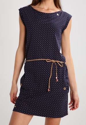 Femme portant une robe sans manches bleu marine avec de petits pois blancs et roses, une ceinture en corde nouée à la taille, et des boutons décoratifs près des poches.
