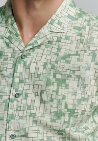 Kortebloemig shirt van lichte stof, met een groen en wit geometrisch patroon, voorzien van een camp collar en turquoise knopen.