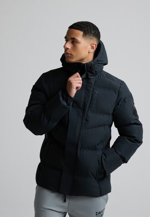 Junger Mann in schwarzer Steppjacke mit Kapuze, der den Reißverschluss anpasst, trägt hellgraue Hose vor einfarbigem grauem Hintergrund.