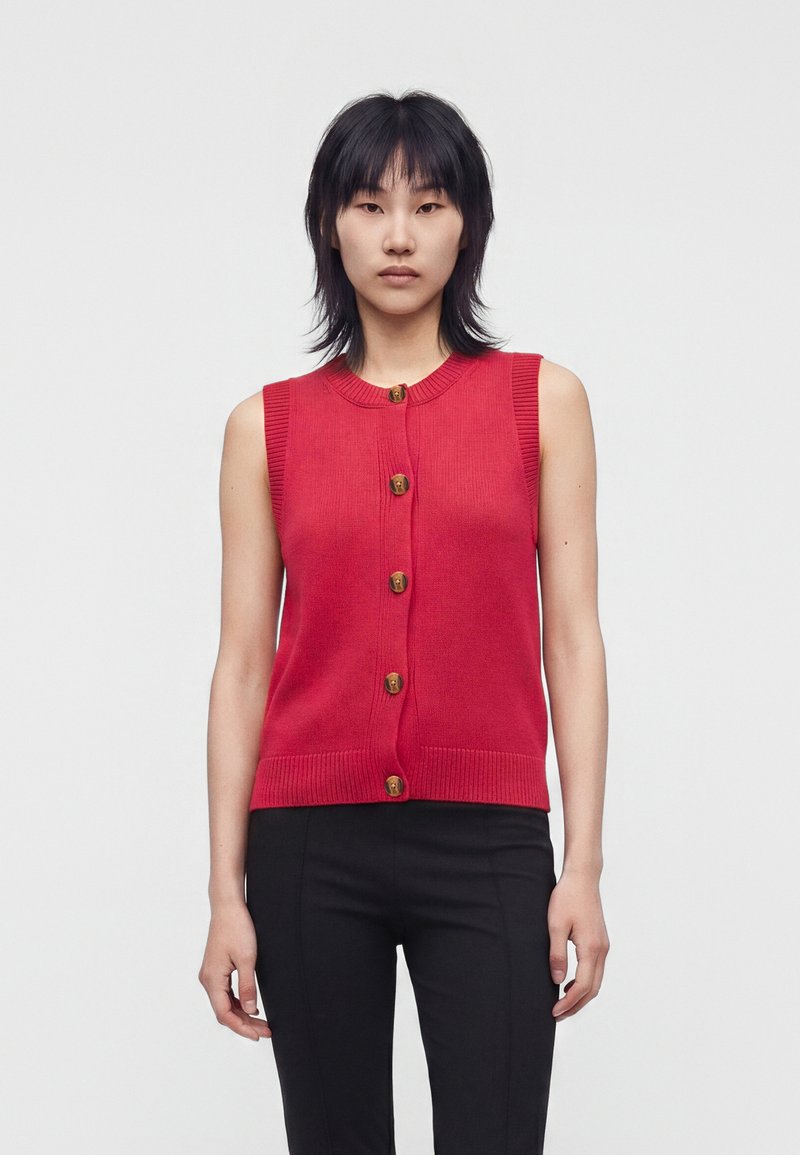 Femme aux cheveux noirs et raides, portant un pull sans manches rouge à boutons et un pantalon noir, debout devant un fond clair uni.