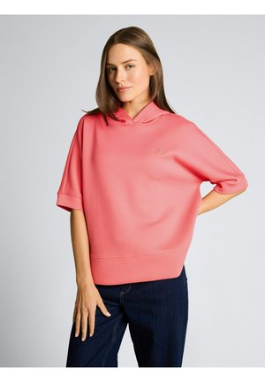 Junge Frau trägt einen lockeren rosa Hoodie mit kurzen Ärmeln, steht mit einer Hand in der Hüfte und der anderen auf dunkelblauer Jeans.