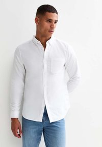Witte lange mouwen knopen-overhemd met een kraag, voorzien van een borstzak, gedragen met blauwe jeans tegen een effen achtergrond.