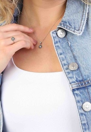 Kvinde iført et sølv solstråle-halskæde og matchende ring, klædt i hvid top og lysblå denimjakke med metalknapper.