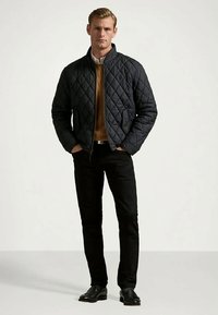 Veste matelassée bleu foncé avec un col montant, fermeture éclair et poches latérales, portée sur un pull marron et assortie à un pantalon et des chaussures noires.