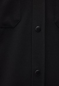 Gros plan sur un tissu noir texturé avec deux boutons-pression noirs et vue partielle de deux poches inclinées sur un vêtement.