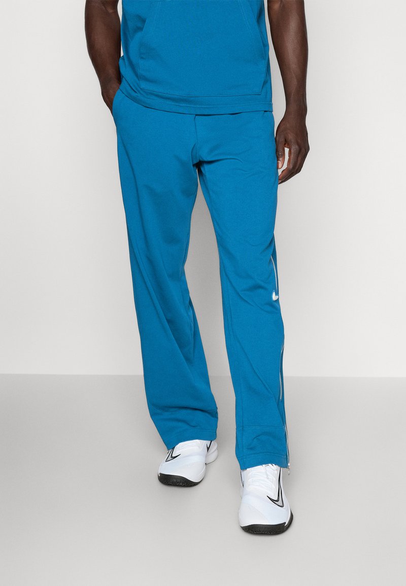 Pantalons de sport turquoise en tissu léger, avec une coupe décontractée et des détails de fermeture éclair sur les côtés, associés à des baskets blanches.