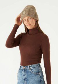 Brown ribbed turtleneck top paired with light blue high-waisted jeans. Worn with a beige knit beanie featuring a logo patch.