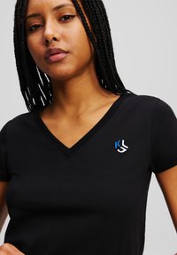 Tricou negru, cu mânecă scurtă, cu decolteu în V. Prezentând un mic slogan albastru și alb în zona stângă a pieptului. Textură de țesătură moale.