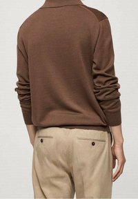 Brauner Strickpullover mit stehendem Kragen und strukturierten Schulterakzenten, kombiniert mit beigen Hosen, die über zwei Gesäßtaschen verfügen.