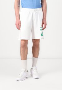 Valged puuvillased šortsid rohelise trükitekstiga ja logoga, millel on paelaga vöökoht ja külgedel lõhed. Kandas valgete tenniste ja heledate sokkidega.