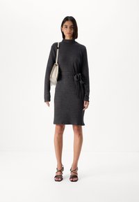 Grå strikket kjole med lange ermer, høy hals og belted midje. Kombinert med svarte stroppesandaler og en lysfarget håndveske.