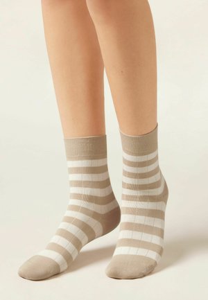 Piedi con indosso calzini alla caviglia a righe orizzontali beige e bianche, in piedi contro uno sfondo chiaro neutro.