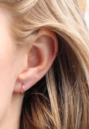 Rosegold-Hoop-Ohrringe, sichtbar am Ohr mit heller Hautfarbe, im Kontrast zu glattem, braun-blondem Haar. Einfaches kreisförmiges Design mit glänzender Oberfläche.