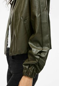 Olivegrüne Kunstlederjacke mit lockerem Schnitt, Druckknöpfen und puffigen Ärmeln. Mit Seitentaschen und samtiger Oberfläche.