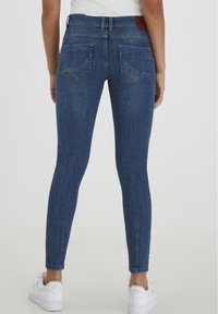 Jean skinny en denim bleu foncé avec une texture lisse, taille haute et deux poches arrière. Présente un motif légèrement délavé et des zips aux chevilles.