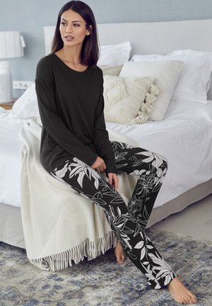 Schwarzes langärmeliges Oberteil und enganliegende schwarze Hose mit weißen Blumenmustern. Umgebung mit einem Bett, einer beigen Decke und einem blau gemusterten Teppich.