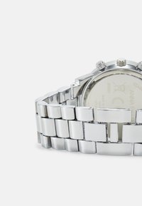 Silverfärgad rostfri stålarmbandsklocka med ett segmenterat armband, rund klockkåpa och tre kronografknappar. Slät, glänsande yta och graverad baksida.