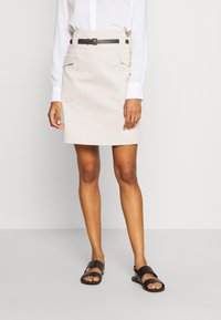 Jupe en coton beige avec des poches latérales et une ceinture noire. Associée à des sandales noires, elle présente une coupe droite et des lignes épurées.