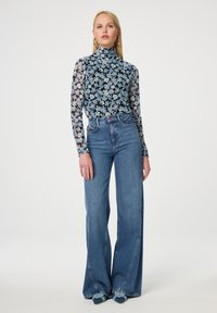 Zwarte mesh coltrui met bloemenpatroon, gecombineerd met lichtblauwe high-waisted, wijde spijkerbroek. Afgewerkt met schoenen met blauwe accenten.
