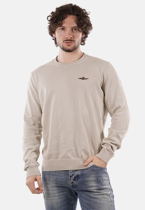 Uomo con capelli ricci che indossa un maglione beige a maniche lunghe con un piccolo emblema sul petto e jeans blu strappati, in piedi con una mano in tasca.