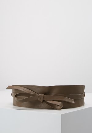 Large ceinture en cuir marron avec un détail de nœud noué, exposée sur un socle d’angle blanc contre un fond clair uni.